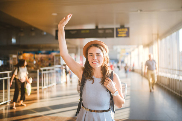 Theme tourism and travel. Young beautiful Caucasian woman in a dress and a hat with a backpack. Tourist mat in the terminal station in long corridor crossing, a schedule board, a gate to raise a hand. - Foto, Imagen