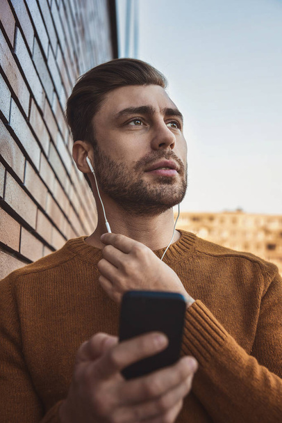 Портрет усміхненого чоловіка з навушниками та мобільним телефоном, що стоїть біля цегляної стіни
. - Фото, зображення