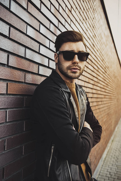 Portret stylowy przystojny młody mężczyzna z włosia, stojący na zewnątrz i opierając się na mur z cegły. Człowiek sobie kurtkę, sweter i okulary przeciwsłoneczne. - Zdjęcie, obraz
