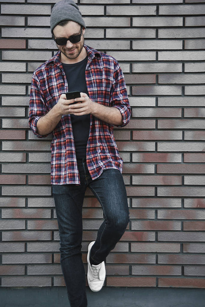 Hipster SMS aplicación de mensajes de texto en la calle de la ciudad en el fondo de la pared de ladrillo. Increíble hombre sosteniendo el teléfono inteligente en la ropa casual inteligente de pie. Estilo de vida profesional joven urbano
. - Foto, imagen