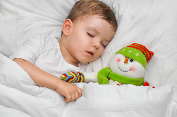 ein süßer Kleinkind-Junge schläft auf weißem Leinen mit seinem Lieblingsspielzeug Schneemann. - Foto, Bild