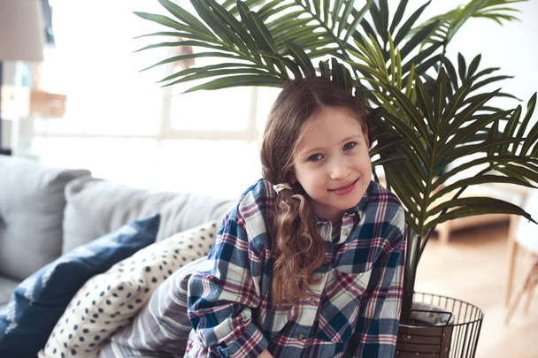happy kid girl sitting on couch near tropical house plant at home - Fotografie, Obrázek