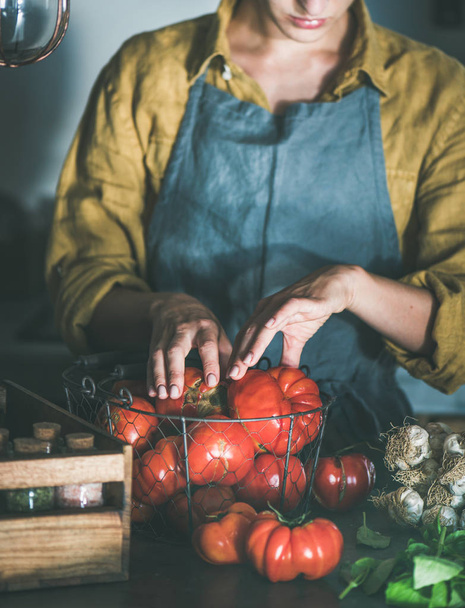 Woman in linen apron taking out from basket ripe heirloom tomatoes for cooking tomato sauce, canned tomatoes or pasta with basil and garlic at kitchen counter. Healthy cooking, slow food or comfort food - Fotoğraf, Görsel