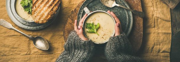 Autumn, Winter home dinner. Flat-lay of Fall warming celery cream soup, grilled bread and female hands over linen tablecloth, top view, wide composition. Comfort food, healthy and slow food concept - Foto, Bild