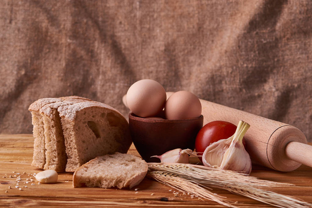 свіжий смачний хліб, концепція випічки. Яйця в глиняній мисці, проростки гарлика, помідорів і пшениці на дерев'яному столі
 - Фото, зображення