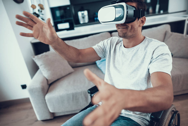 Disabled Man Enjoys Virtual Reality Goggles. Portrait of Handicapped Person Sitting in Wheelchair, Parked in Large Bright Living Room. Male in Casual Clothes Uses VR Headset Keeping Hands Up - Фото, изображение