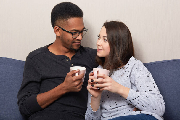Horizontal shot of lovely multiethnic girlfriend and boyfriend drink coffee or tea together, pose at comfortable couch, look at each other with love, enjoy comfort, togetherness. Interracial relations - Foto, Bild