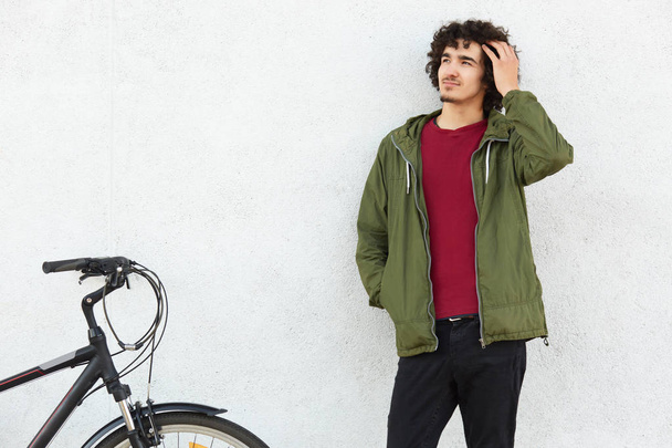 Handsome dreamy male cyclist has curly hair, dressed in casual green anorak, keeps hand in pocket, rides bicycle, concentrated aside, isolated over white concrete wall. People and youth concept - Photo, image