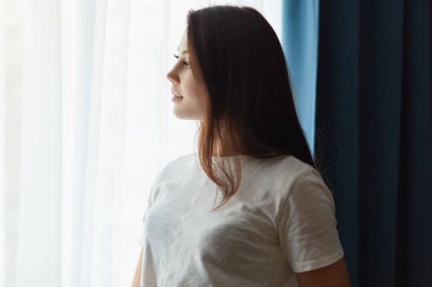Sideways shot of thoughtful dark haired woman dressed in white t shirt, thinks about something while stands near window, enjoys calm atmosphere at home, feels lonely. Contemplative lady indoor - 写真・画像