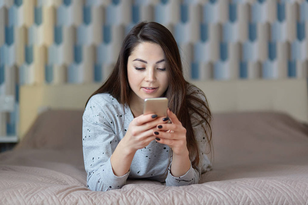 Photo of attractive concentrated woman holds mobile phone, sends messages to friend, downloads music files, connected to high speed internet, poses at comfortable bed alone. Online shopping. - 写真・画像