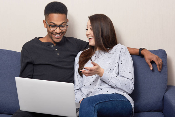 Cheerful mixed race couple use laptop computer for watching movies, sit together on sofa over home interior, browse information, connected to wireless internet, enjoy spare time. Interracial relations - Foto, Bild