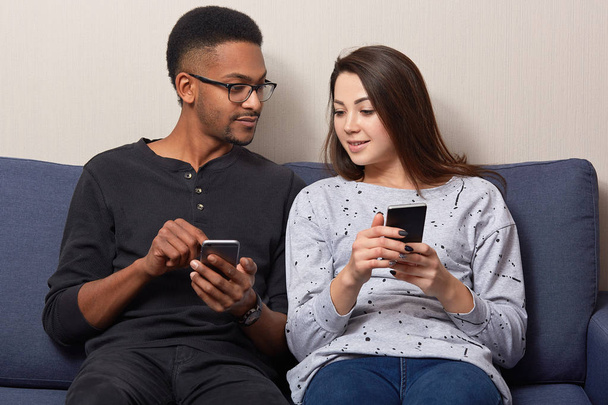 Photo of mixed race people update application on mobile phones, enjoys spare time with modern technologies, holding digital devices, dressed in jumpers and jeans, pose at sofa in their flat. - Φωτογραφία, εικόνα