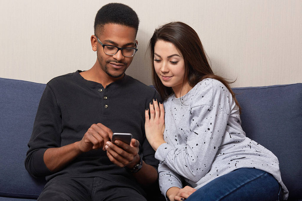 Multiethnic couple reads news in social network or watch movie online on cell phone, sit closely on sofa, concentrated on recieved media file, browse internet online. People, interracial friendship - Foto, afbeelding