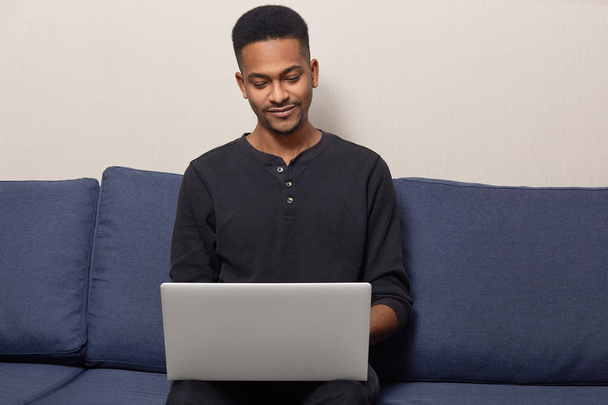 Photo of pleased dark skinned guy has beard, wears black sweater, poses at comfortable sofa, installs application, connected to wireless internet at home, reads information for project work. - Foto, Imagem