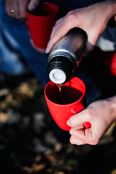 woman hands pours hot tea or coffee out of thermos on winter forest background. girl using a thermos in on a snowy mountain. red cup - Photo, image