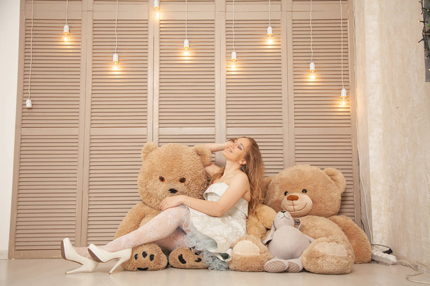 cute caucasian girl in white dress playing with plush brown teddy bears in her room on the background of light bulbs near the wooden closet - Photo, Image