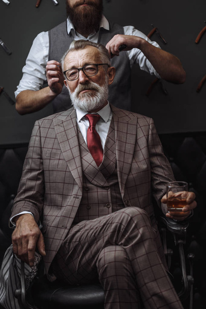 Bearded old businessman sitting at barber shop in chair choosing haircut design - 写真・画像