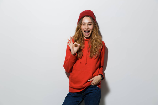 Portrait of a cheerful young woman wearing hoodie standing isolated over white background, showing ok gesture - Фото, зображення