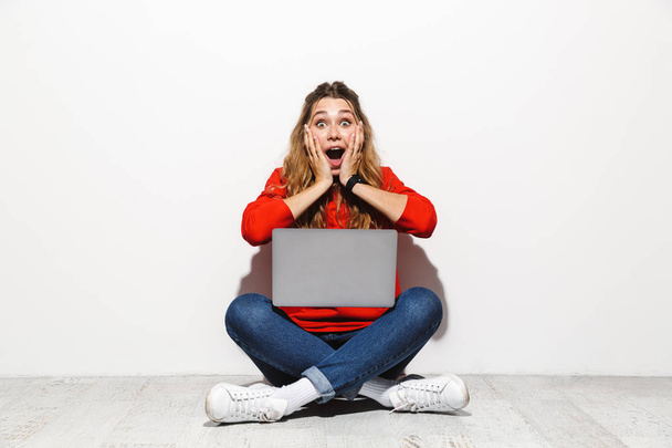 Portrait of an excited young woman wearing hoodie sitting isolated over white background, using laptop computer, celebrating success - Foto, Bild