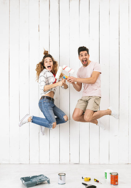 Full length photo of cool couple man and woman jumping while painting white wall and making renovation using paint rollers - Fotografie, Obrázek