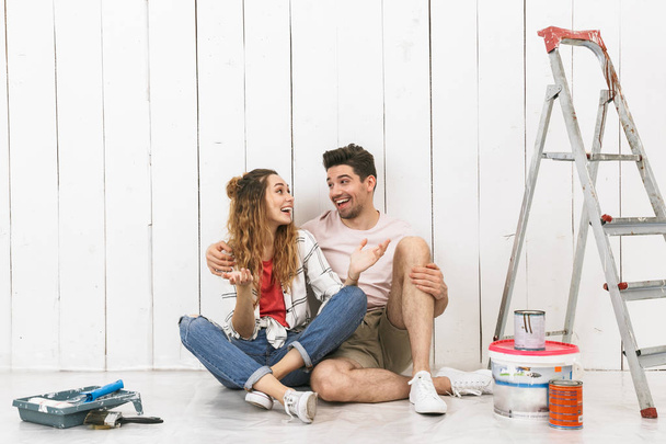 Photo of young couple man and woman sitting on floor over white wall near ladder while making renovation indoor - Foto, afbeelding