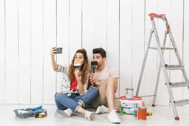 Photo of adorable couple man and woman sitting on floor over white wall near ladder and taking selfie on cell phone while making renovation indoor - Fotó, kép