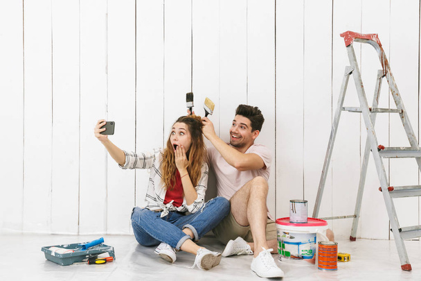 Photo of funny couple man and woman sitting on floor over white wall near ladder and taking selfie on cell phone while making renovation indoor - Fotografie, Obrázek