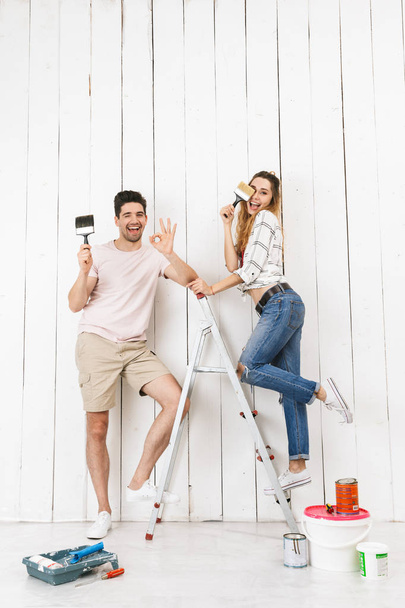 Full length photo of joyous couple man and woman standing on ladder while painting white wall and making renovation indoor - Foto, Bild