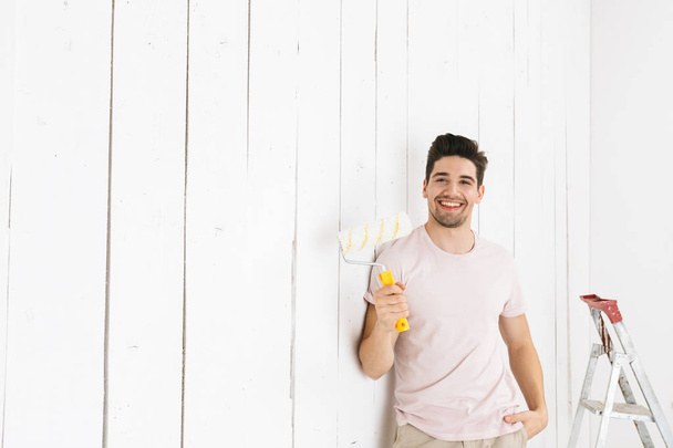 Image of joyful man 20s painting white wall and making renovation using paint roller - Photo, Image