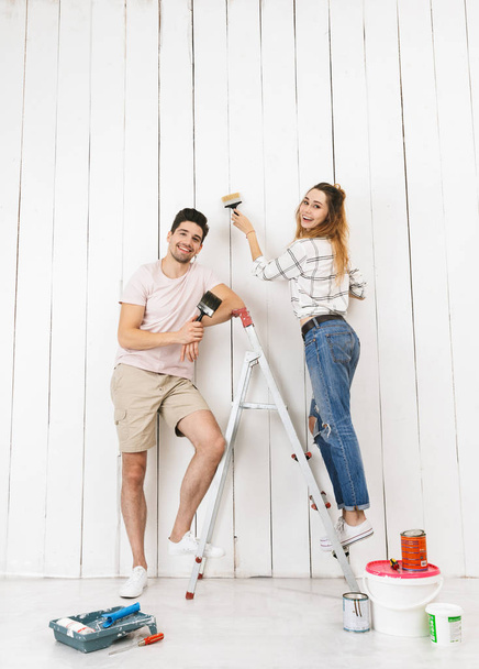 Full length photo of smiling couple man and woman standing on ladder while painting white wall and making renovation indoor - Foto, Imagen