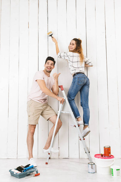 Full length photo of european couple man and woman standing on ladder while painting white wall and making renovation indoor - Foto, Imagen