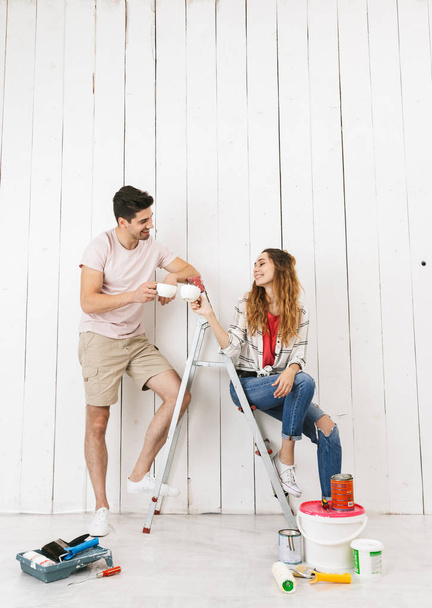Full length photo of cheerful couple man and woman resting on ladder after painting white wall and making renovation indoor - Foto, Imagen