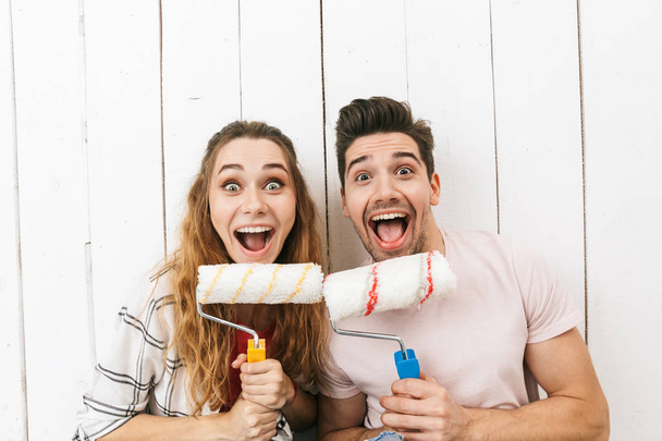 Image of funny couple man and woman holding paint rollers and standing over white wall while painting and making renovation - Fotografie, Obrázek