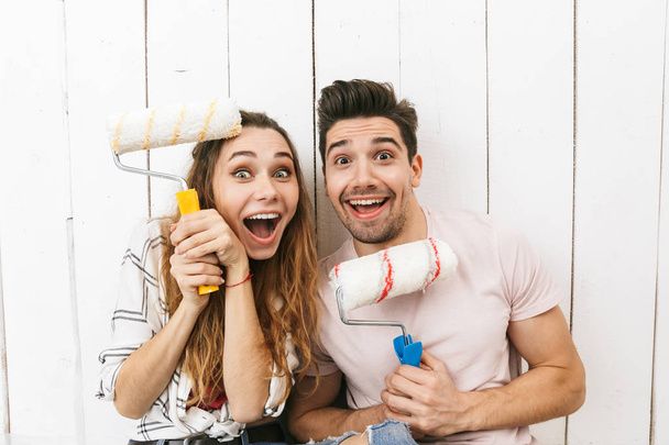 Image of joyful couple man and woman holding paint rollers and standing over white wall while painting and making renovation - Foto, Imagem