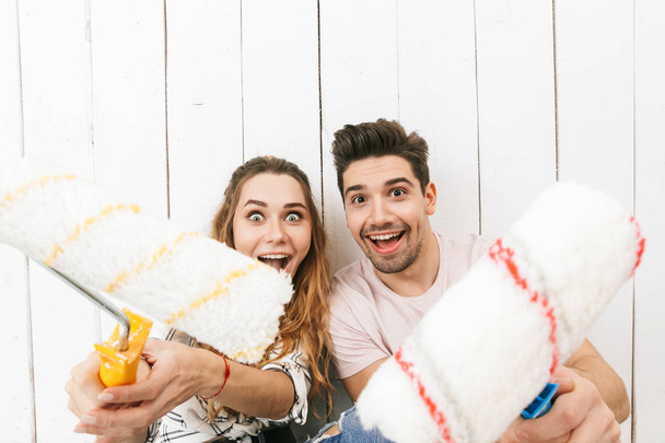 Image of european couple man and woman holding paint rollers and standing over white wall while painting and making renovation - Фото, изображение