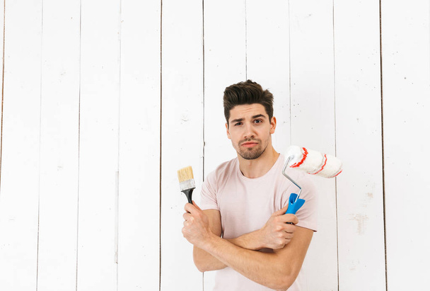 Portrait of young man 20s holding paint roller and brush while painting white wall and making renovation - 写真・画像