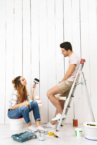 Portrait of attractive couple man and woman using ladder while painting wall and making renovation indoor - Foto, Bild