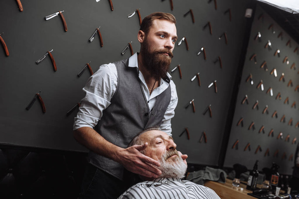 Male barber chooses beard design for old-aged client at hairdresser. - Foto, afbeelding
