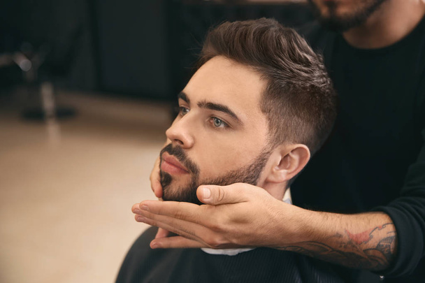 Hairdresser applying serum onto client's beard in barbershop. Professional shaving service - Photo, image