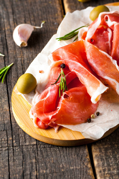 Closeup of thin slices of prosciutto with mixed olives and paprika on wooden cutting board - Fotó, kép
