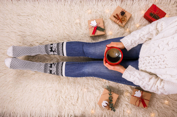 junge Frau, die Kaffee trinkt und auf dem pelzigen Teppich sitzt. Nahaufnahme weiblicher Beine in warmen Socken mit Hirschprint, Packpapierrollen, Draufsicht. Weihnachtsvorbereitungskonzept. Hintergrund, Kopierraum - Foto, Bild