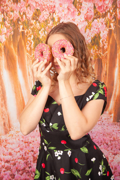 schönes junges Mädchen steht in schwarzem Pin-Up-Kleid und hat Spaß beim Spielen mit glasierten rosa Donuts - Foto, Bild