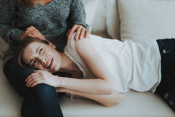 Upset Young Daughter Lying on Legs of Mother on Sofa. Relationship in Family. Worried Mother. Psychological Help. Unhappy Girl. Support Concept. Scared Person. Women on White Sofa. - 写真・画像