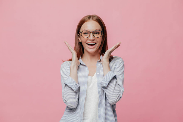 Studio shot of joyful excited woman dressed in striped shirt, spreads hands near face, wears optical glasses, models over pink background, doesnt expect to notice something pleasant and unexpectable - Photo, Image