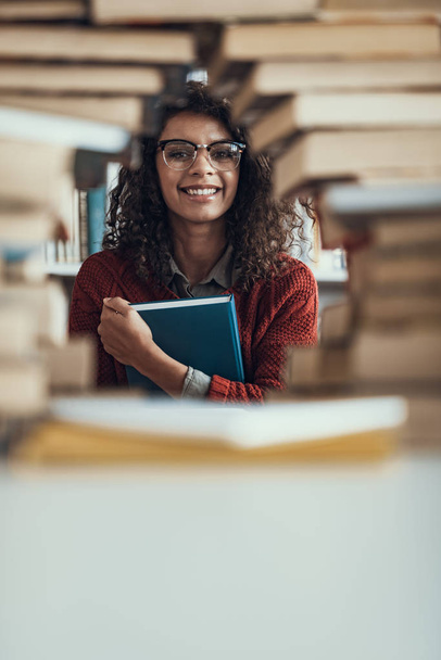 Positive student looking through the pile of books and smiling - Фото, изображение