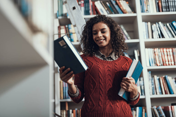 Young lady looking glad while choosing books in the library - Foto, afbeelding