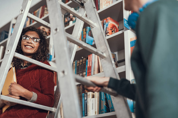 Cheerful emotional curly student pressing a book to her chest while sitting on the step ladder and smiling - Photo, image