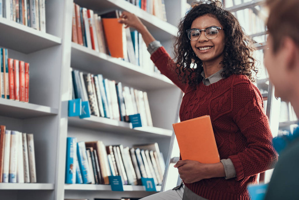 Pretty curly lady smiling and taking books from the shelf - Foto, imagen