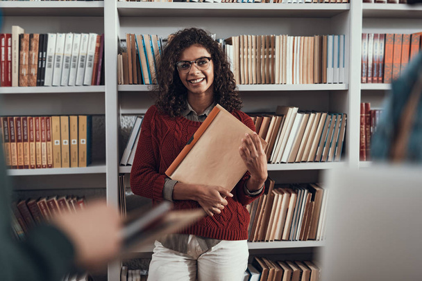 Waist up of cheerful lady standing with folders near the bookcase - Foto, Bild