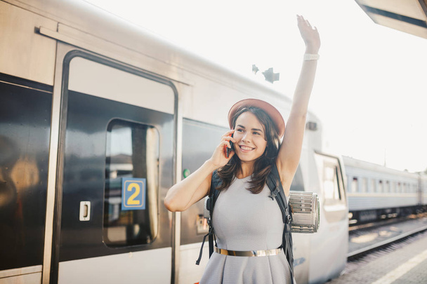 Theme transportation and travel. Portrait young caucasian woman with toothy smile standing train station train background with backpack using technology, smart phone hands in dress and hat summer. - Photo, Image